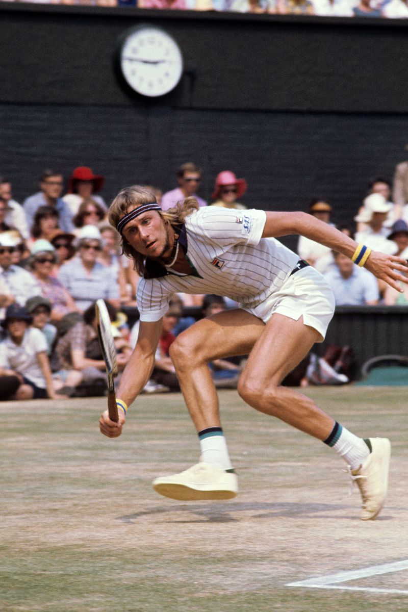 Tennis player Bjorn Borg on the court hitting a ball. He wears short white shorts and a tight-fitting white pinstriped polo. He wears his hair long with a headband around his forehead.