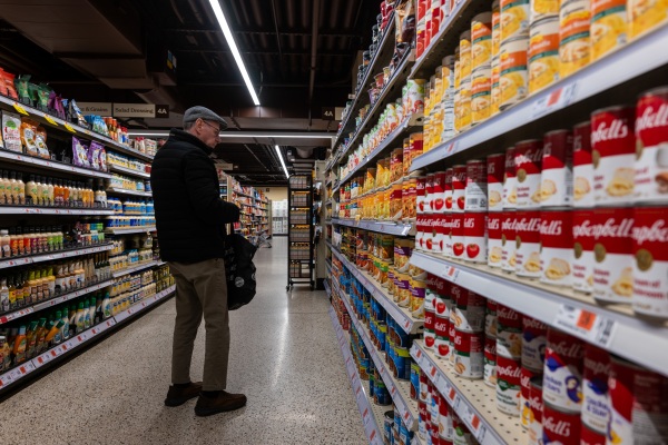 This photo shows cans of soup, vegetables and beans lining the shelves of a grocery store aisle. A man stands in the aisle, looking at the cans.