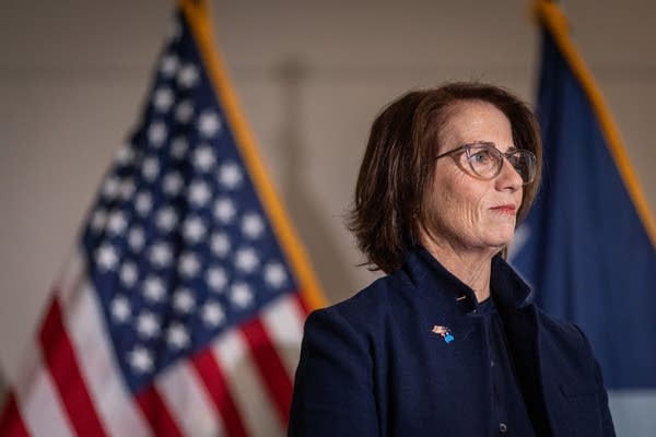 A woman stands in front of a U.S. and Minnesota state flag