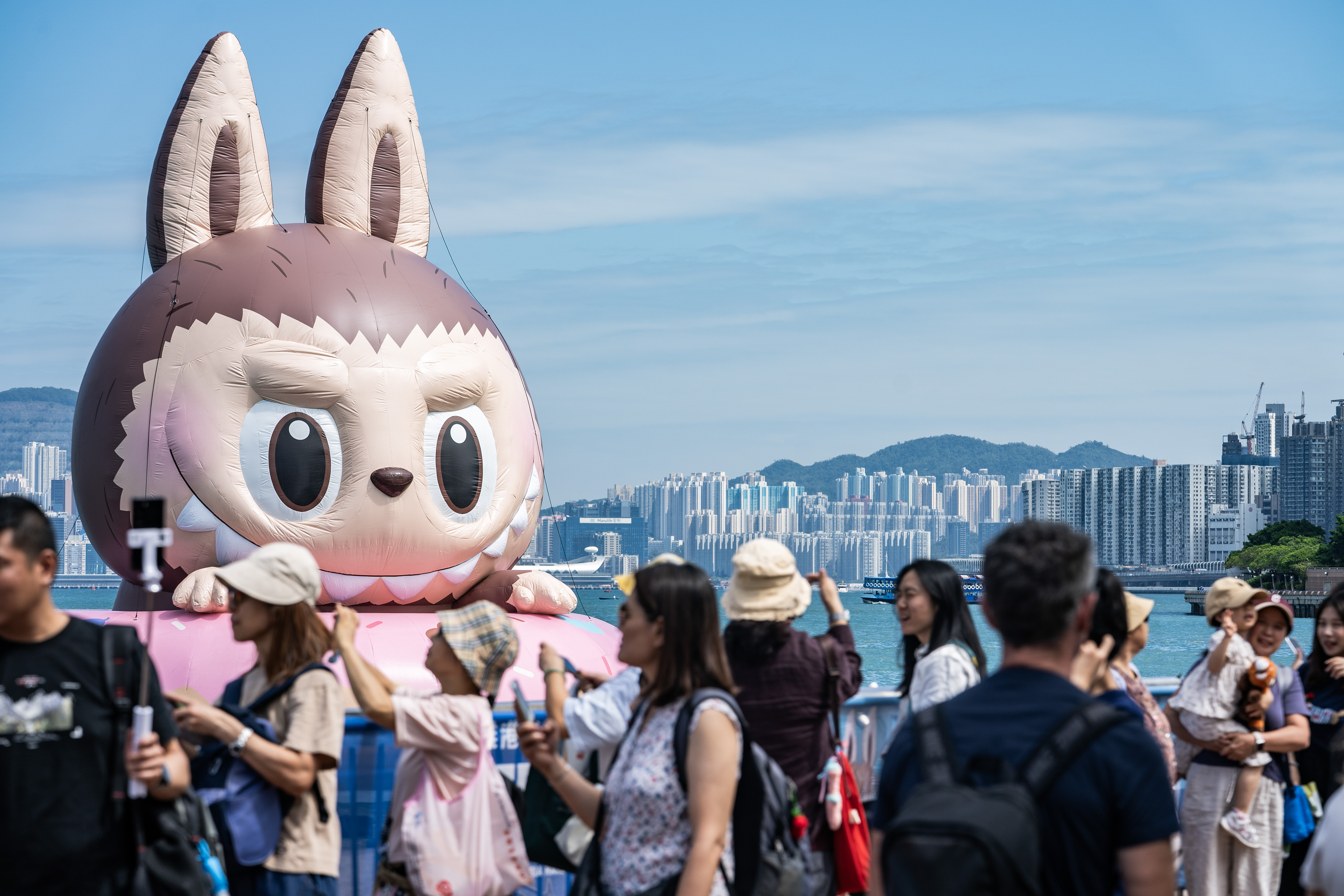 A huge inflatable Labubu in the harbor of Hong Kong, with people milling about in the foreground.