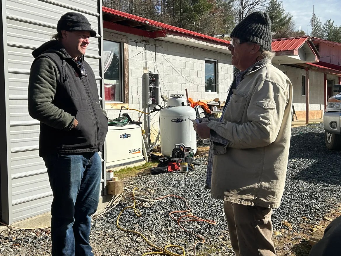 Two men wearing jackets and hats speak on a gravel road in front of a grey and red building.