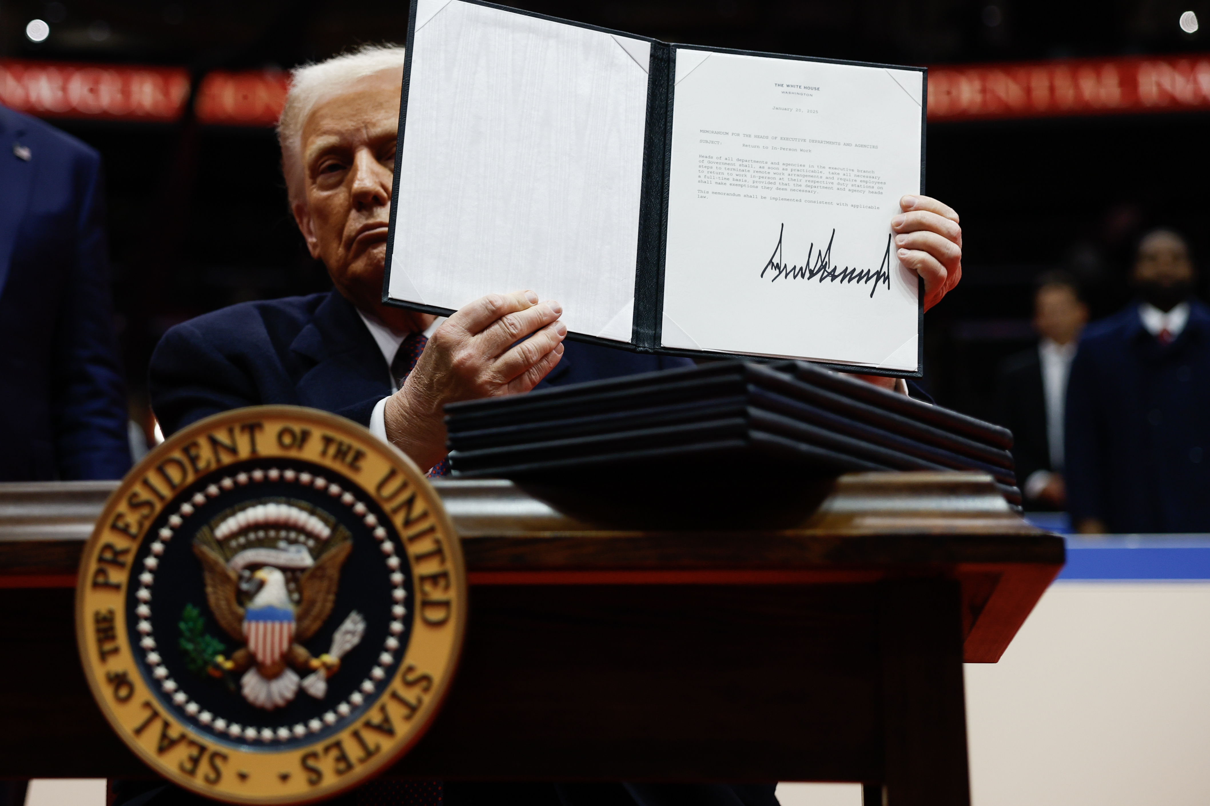 President Trump, standing at a podium, holds up an executive order with his signature very large at the bottom