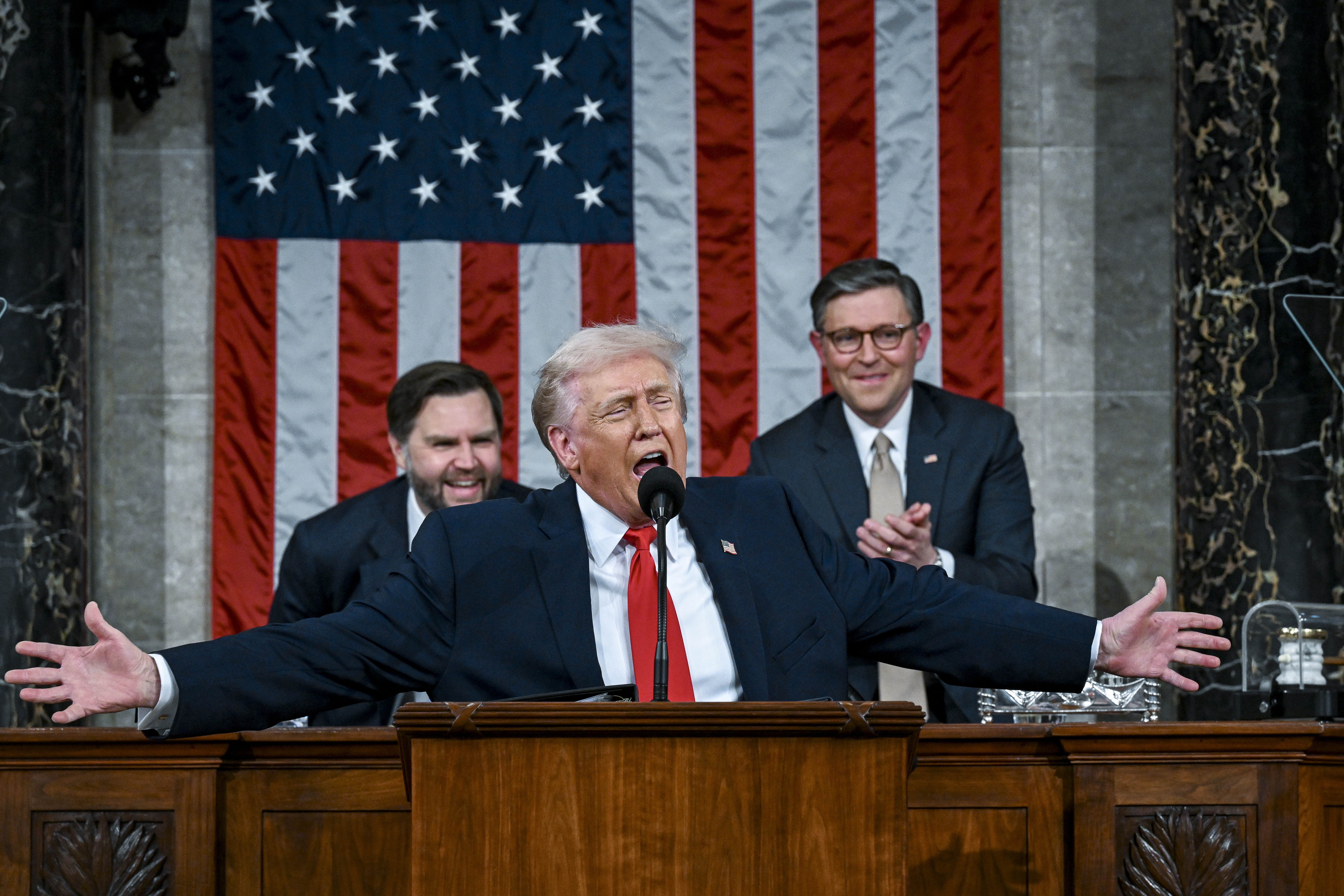 Donald Trump, his arms spread, speaks into a microphone; behind him is an American flag, and Vice President JD Vance (L) and House Speaker Mike Johnson (R) are visible over either shoulder.
