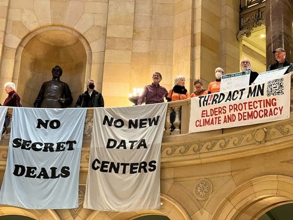 People on the balcony of the Minnesota Capitol hang signs protesting data centers.