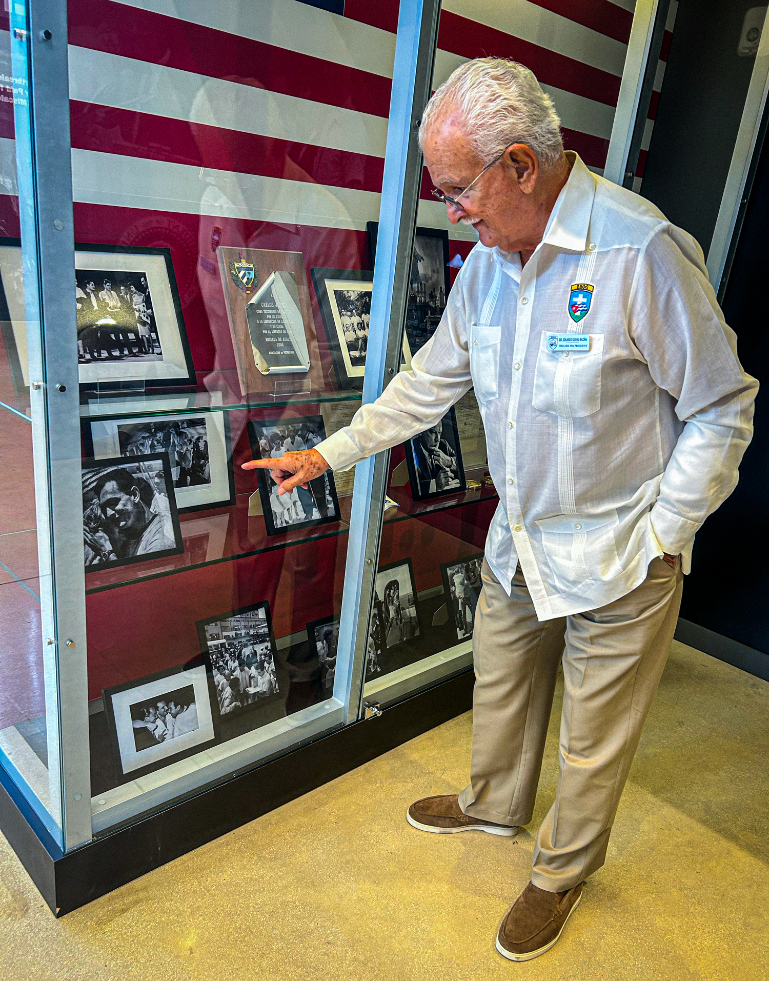 An elderly man point to framed black-and-white photos held in a glass display case.