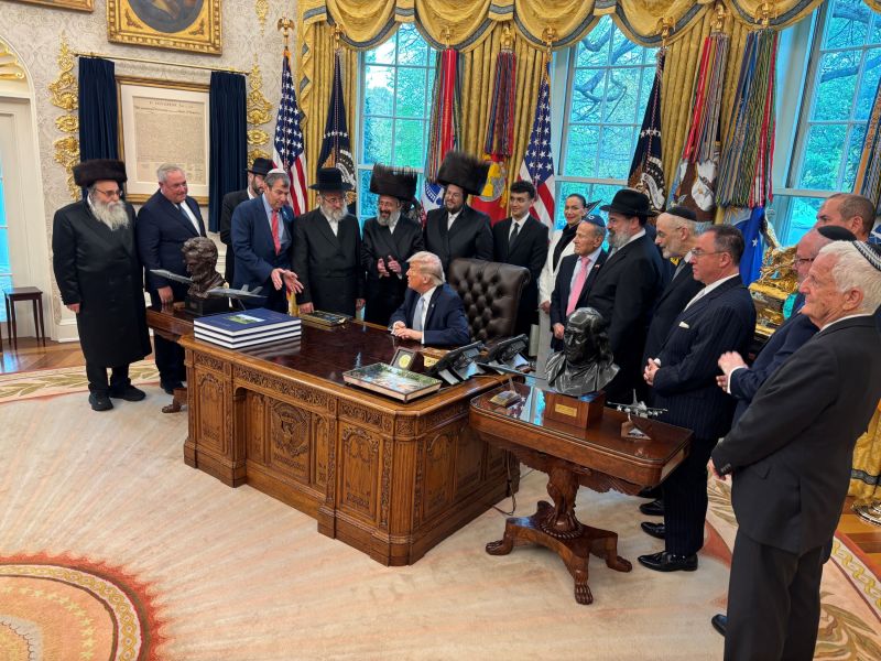One woman and over a dozen men, most in headwear associated with Jewish religious practice, stand around President Trump in the Oval Office. He is seated at a desk.