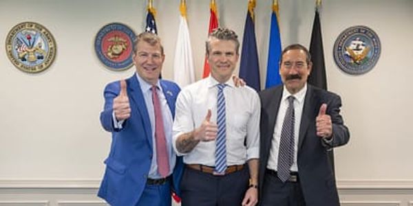 A photograph shows three white men in dress clothes standing together, giving thumbs up. They are in front of flags and wall medallions representing US military branches.