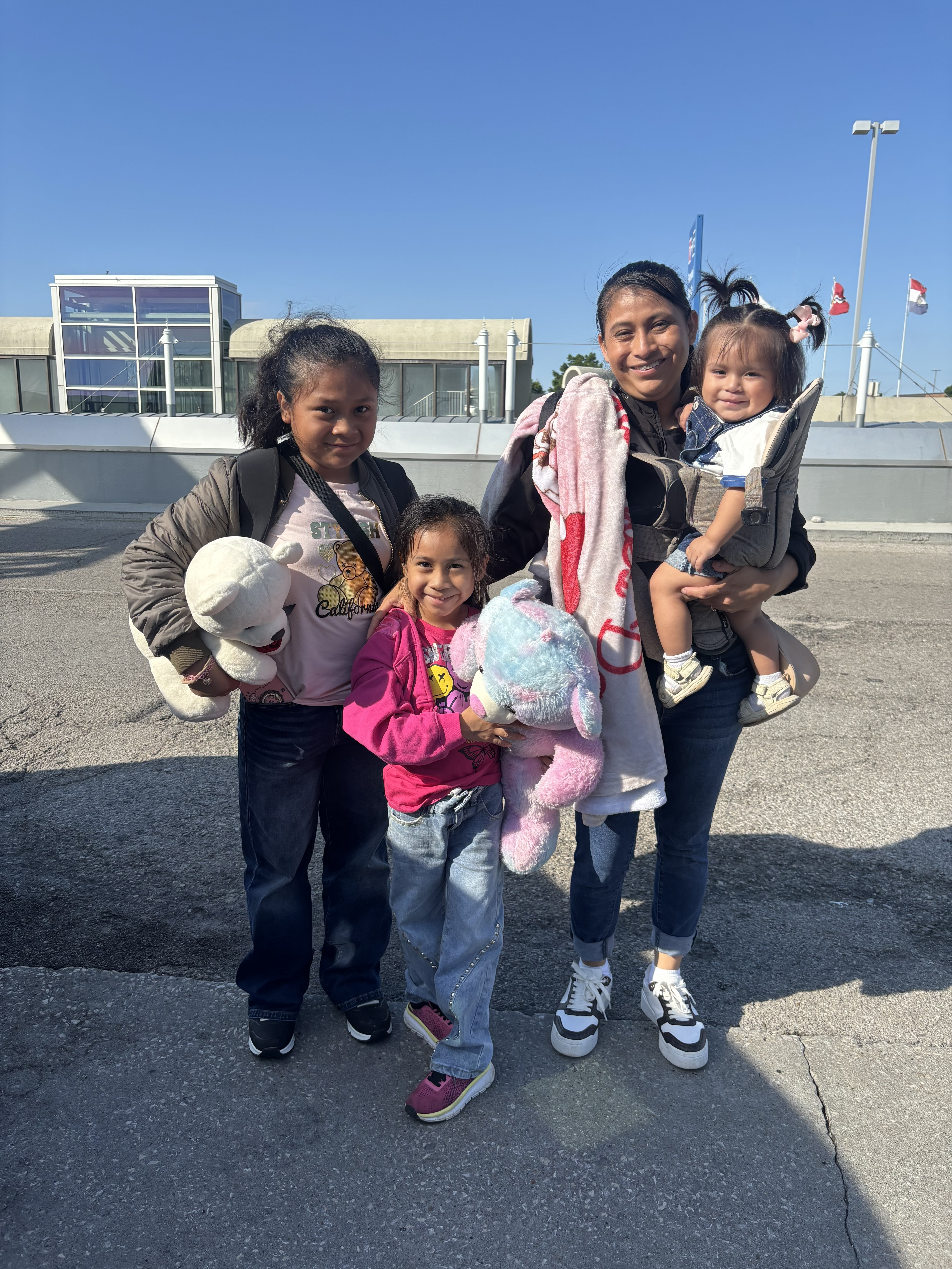 A young Guatemalan woman holds her toddler daughter in a sling, standing alongside her two other young daughters at an airport drop-off.