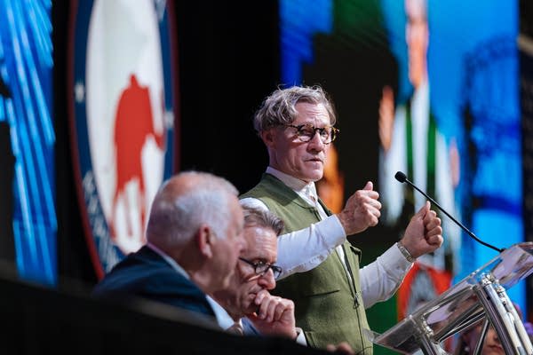 A man in a green vest speaks at a podium.