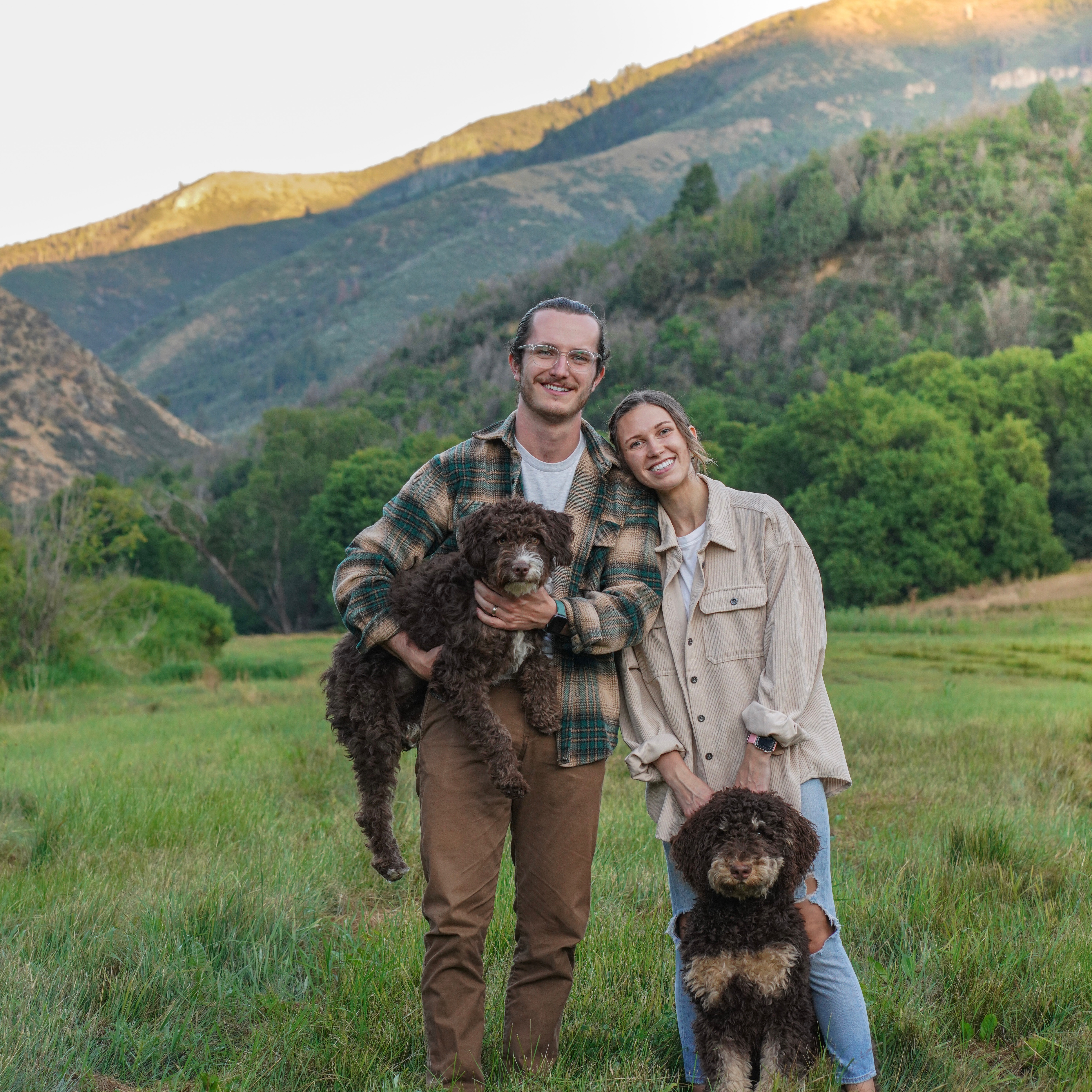 A smiling man and woman stand in a grassy meadow with forested mountains in the background. The man holds a brown, curly-haired dog in his arms, while a second dog sits at the woman's feet.