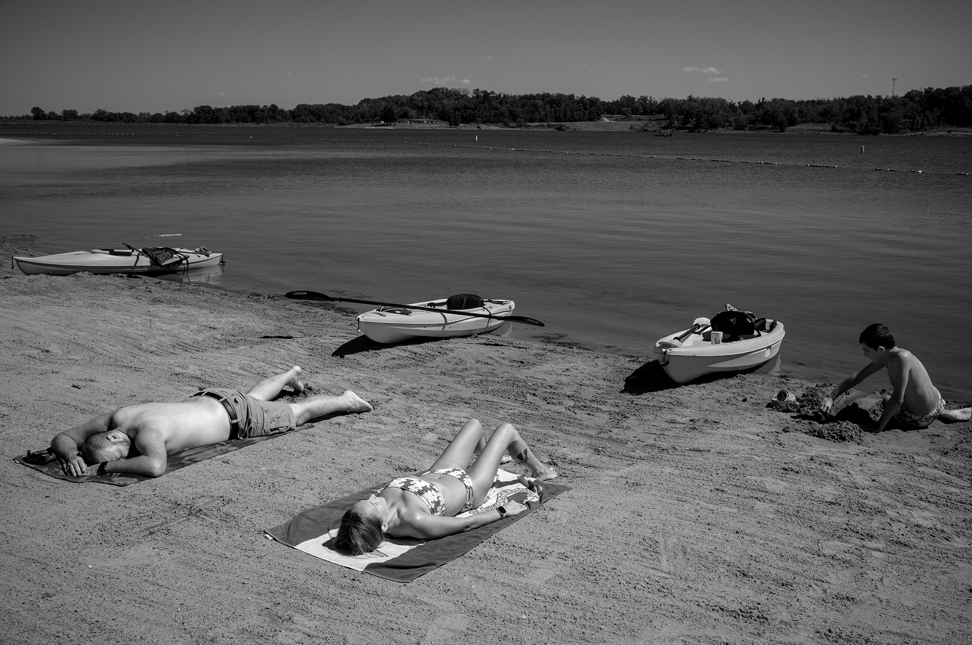 Two people laying on a beach with kayaks nearby. Another person plays in the sand.
