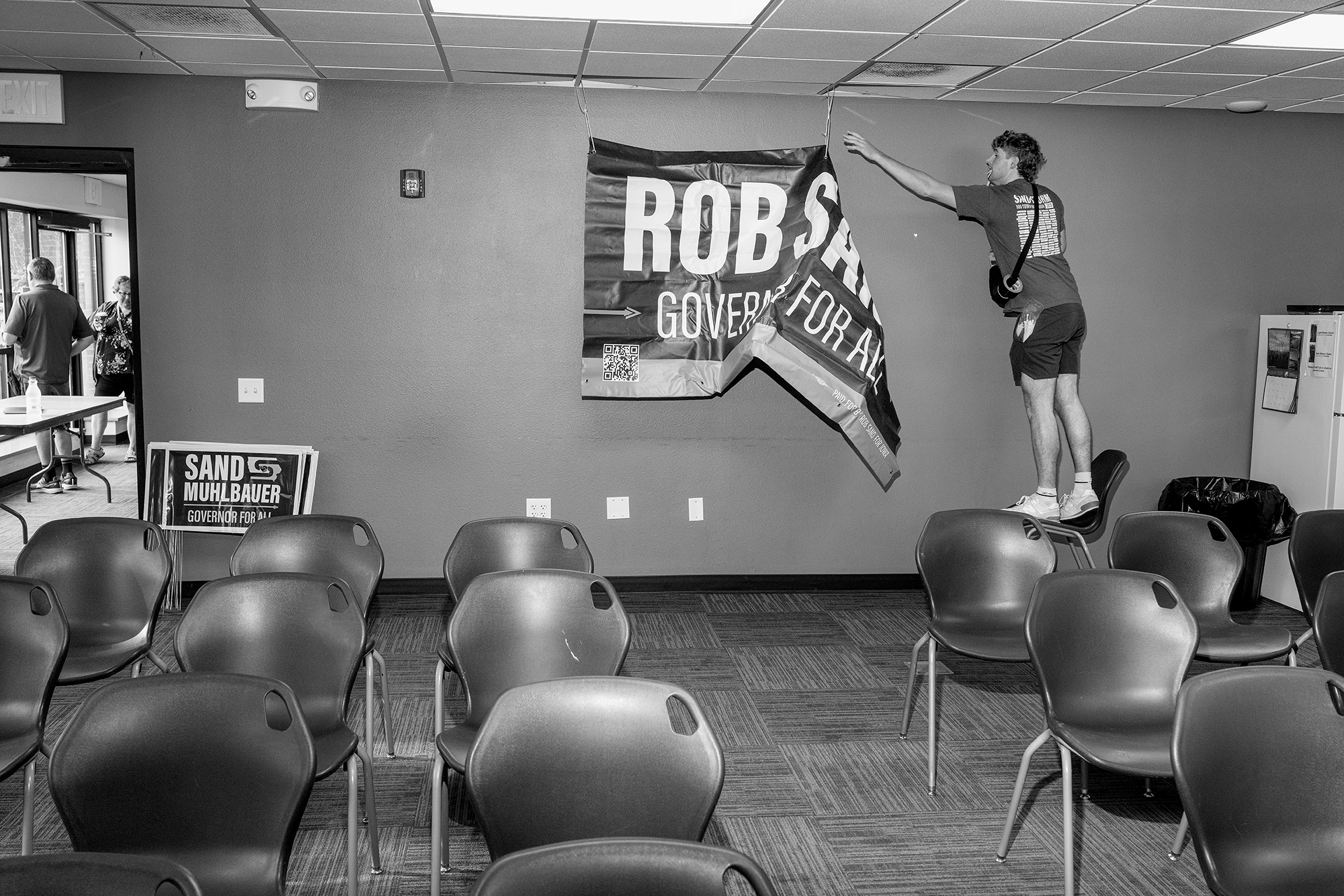 Man stands on a chair, taking down a "Rob Sand for Governor" sign in a conference room.