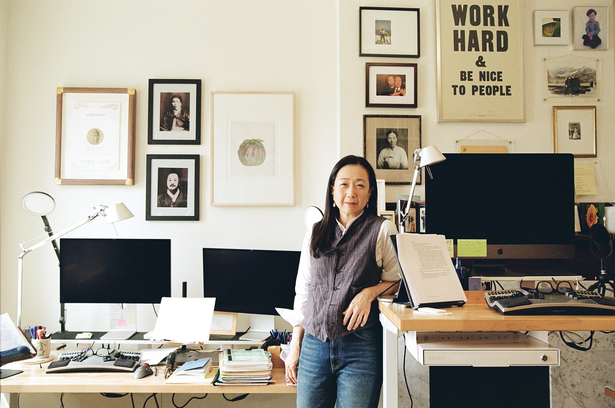 A middle-aged Korean woman stands with her elbow propped on a standing-height desk, surrounded by computers and photos on the walls.