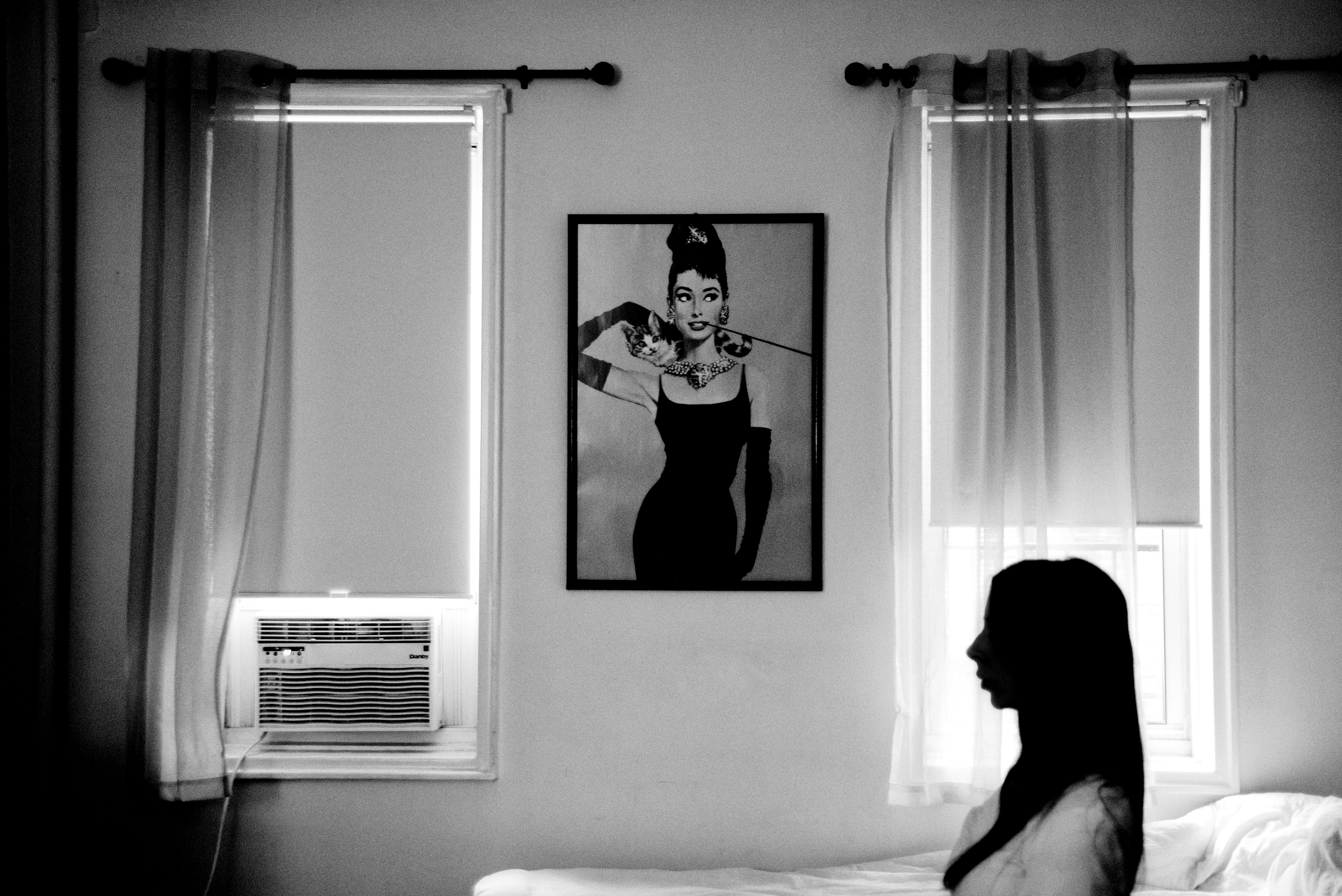 A woman sits silhouetted in a bedroom in which a photo of Audrey Hepburn hangs on the wall.