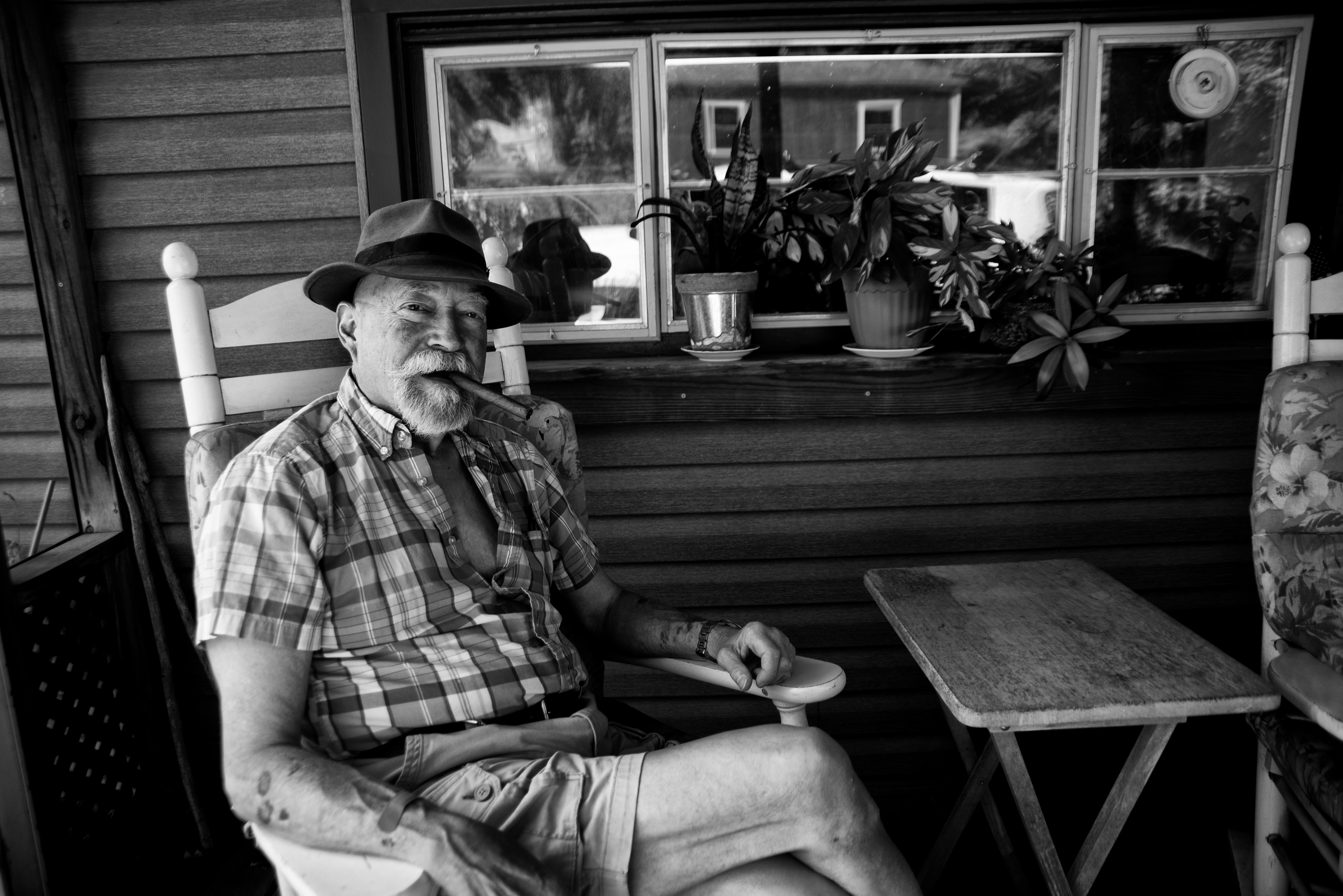 A man wearing a hat sits on a front porch next to a small table.