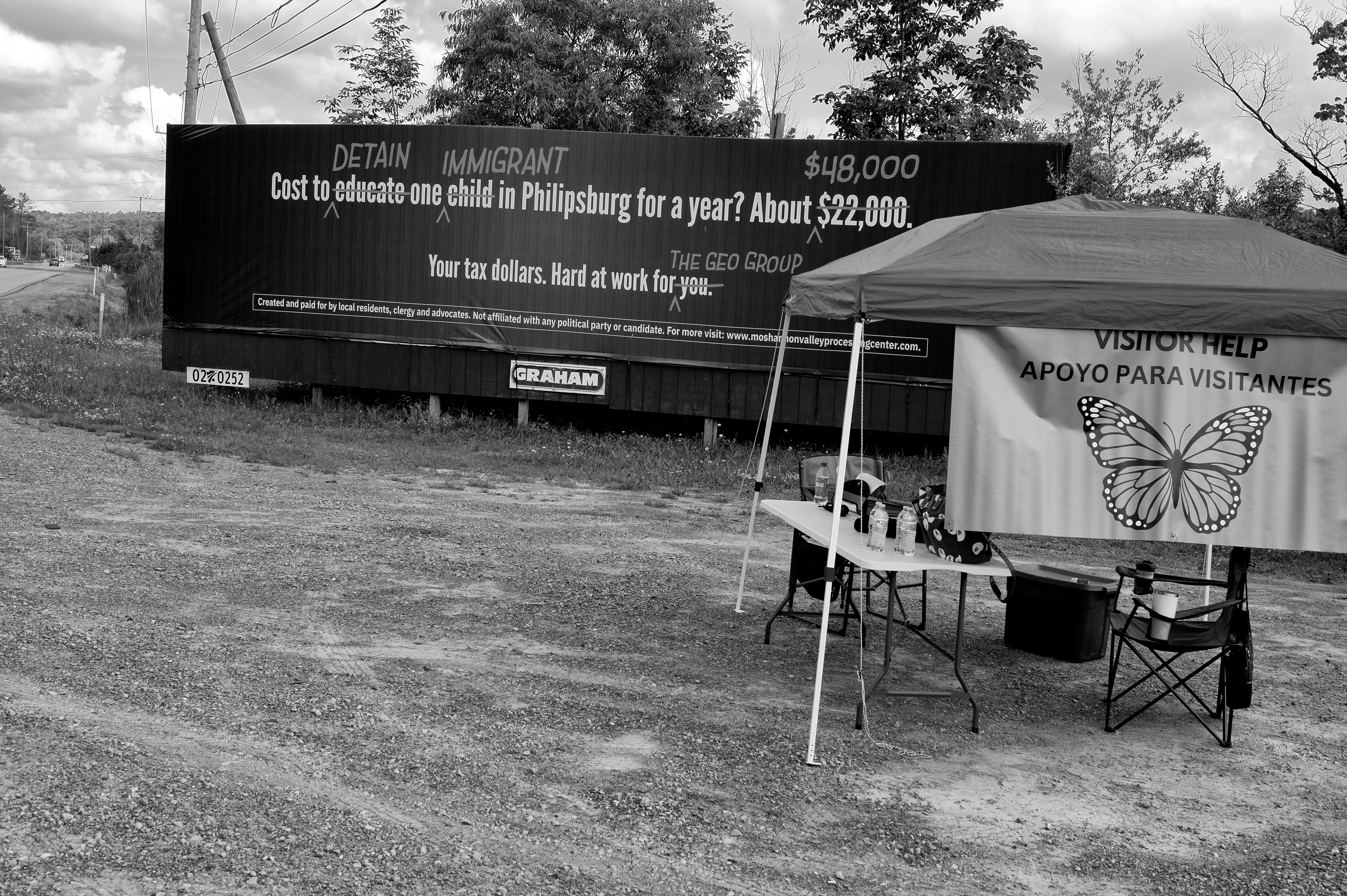 A tent with a table sits in front of a billboard criticizing ICE detentions.