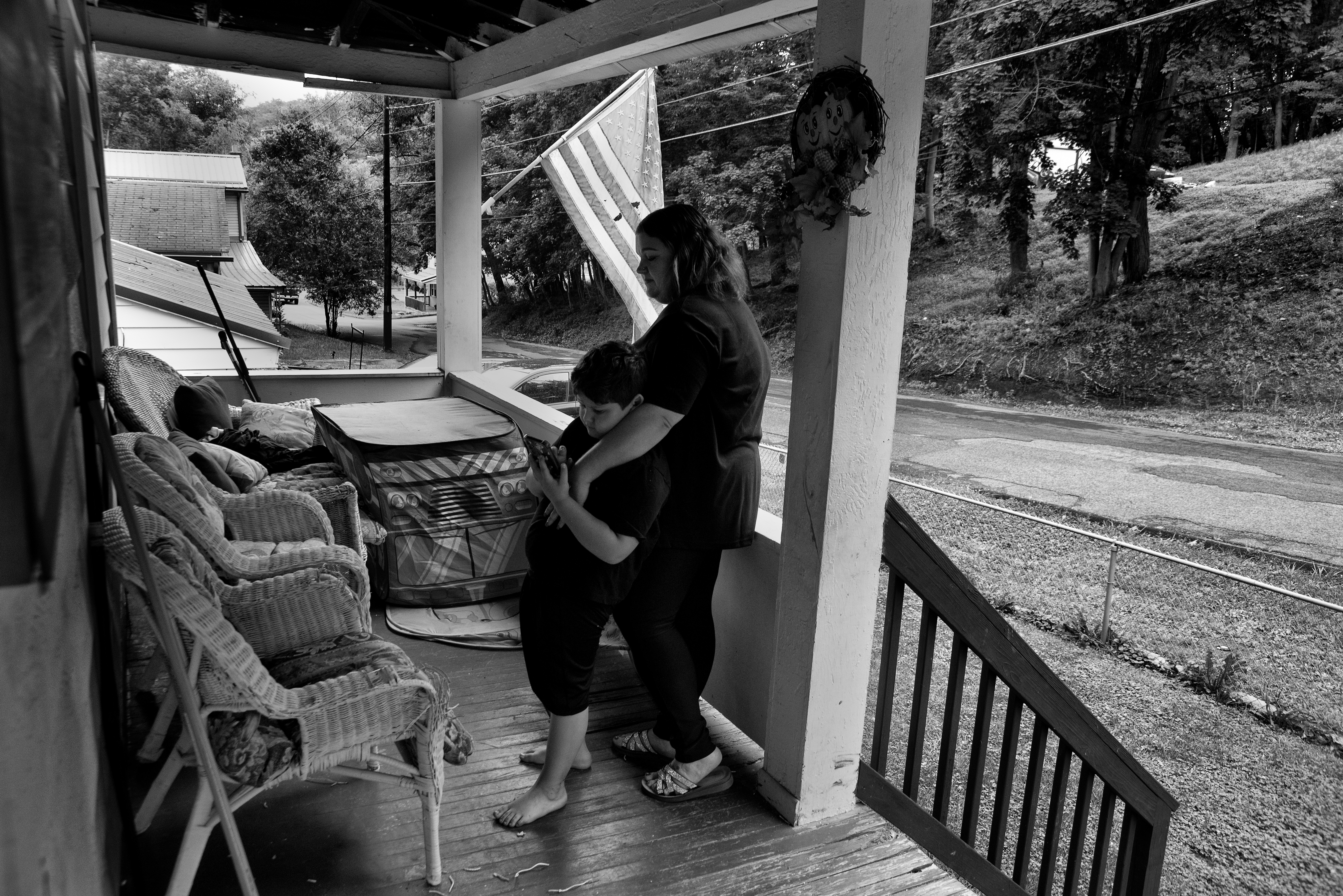 A mother and son stand on the front porch of a house with an American flag flying out front.