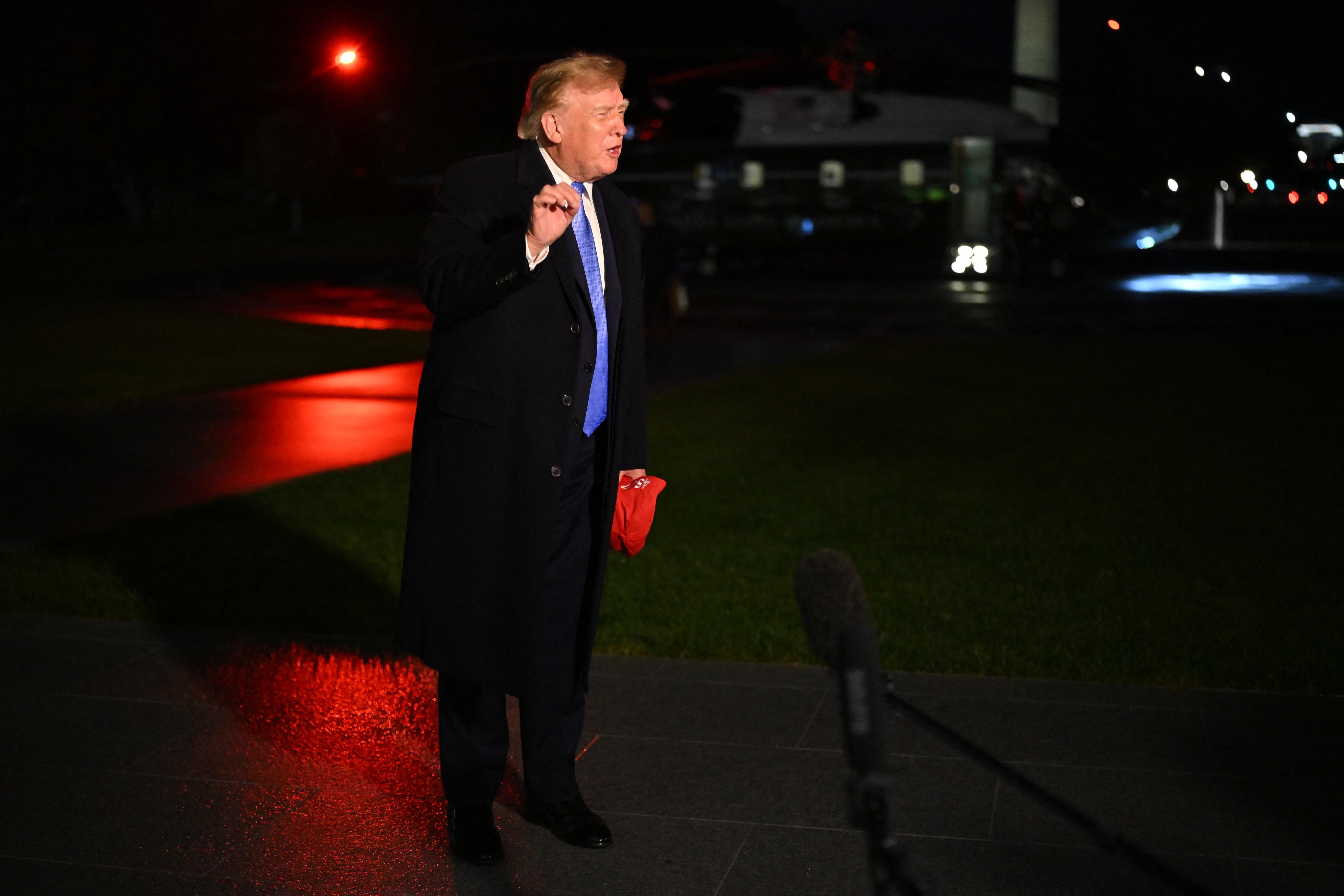 Donald Trump, wearing a dark topcoat over a suit and holding a red hat in one hand, gestures with the other as he speaks in front of the Marine One helicopter at night.