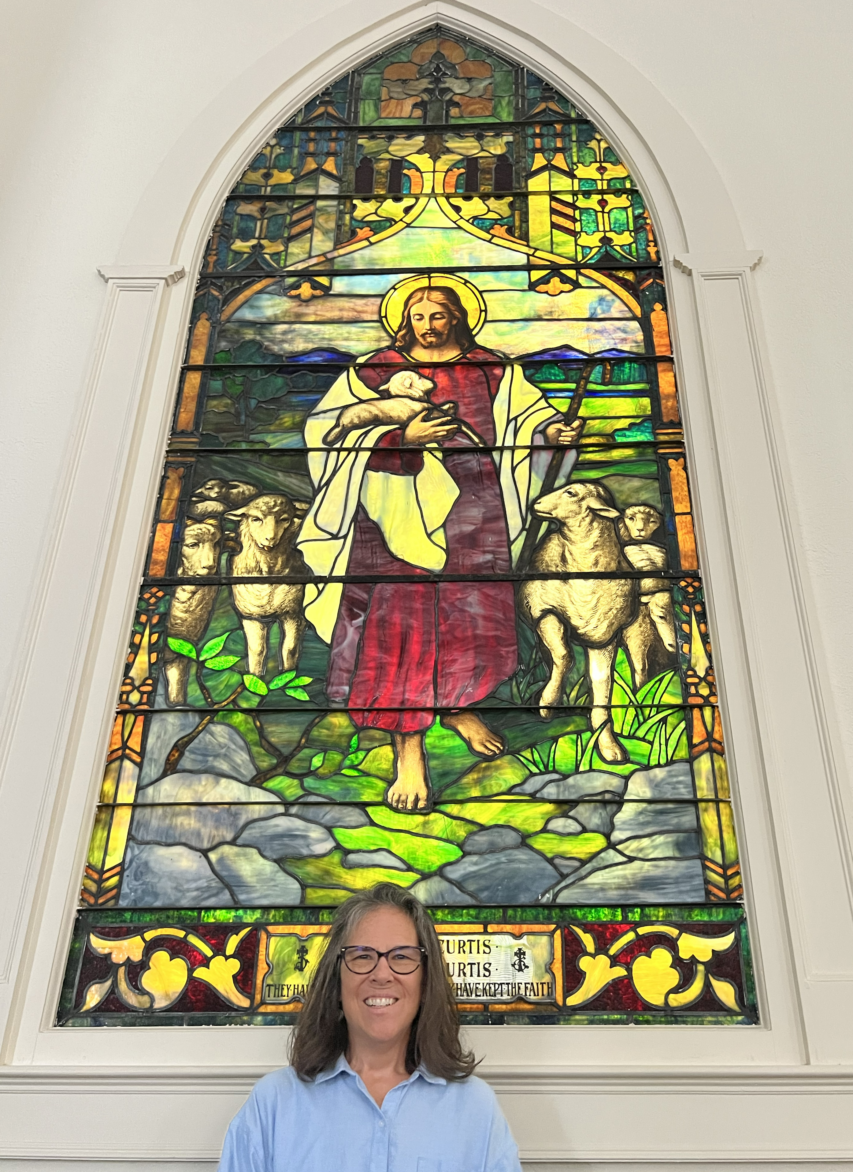 A bespectacled middle aged woman smiles in front of a stained glass window depicting Jesus as a shepherd.