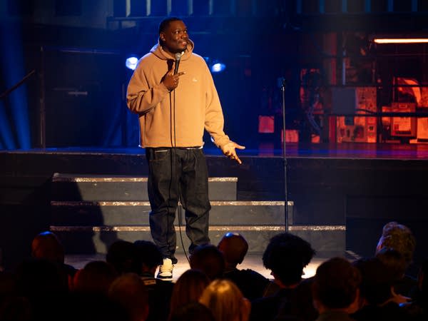 A comedian holds a microphone while performing on a stage.