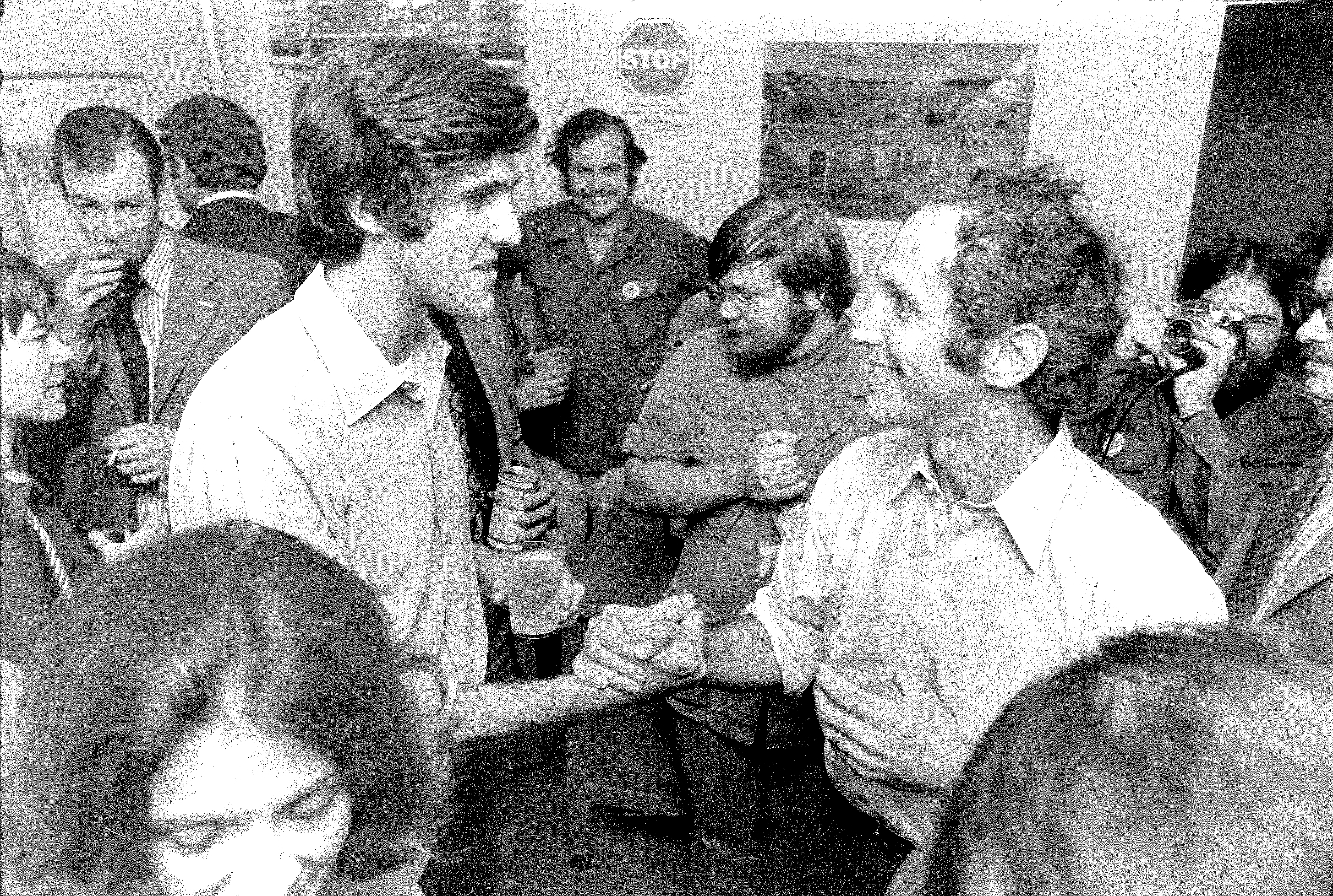 In this black and white photo at a reception, a very young John Kerry and Ellsberg, hands clasped in a thumbs-up handshake, and both wearing white button-up shirts, smile at one another as others mill about drinking cans of Budweiser.