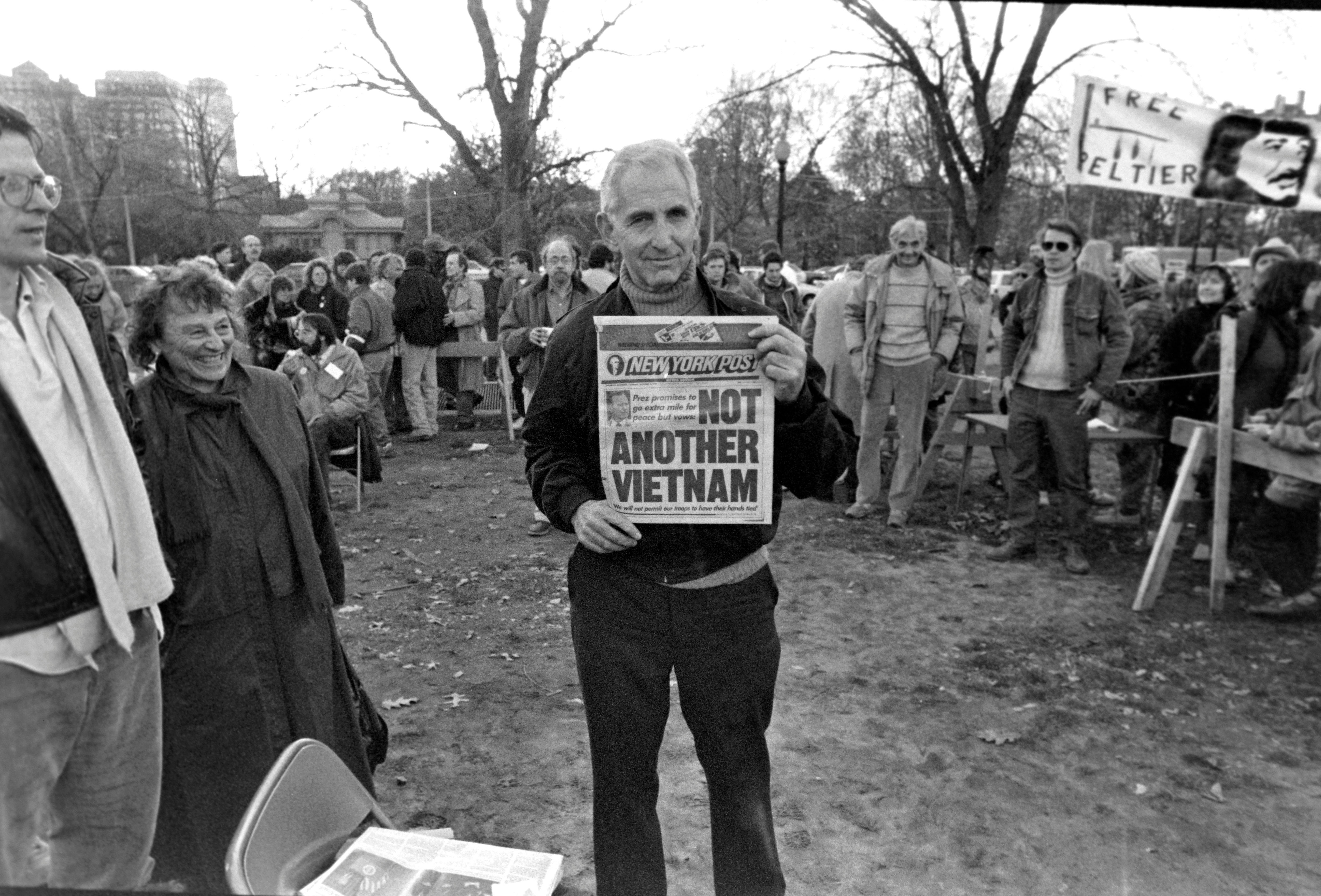 Ellsberg, hair now gray and dressed in dark casual clothes, stands in a grassy clearing surrounded by people, and some signs, holding a copy of the NY post. Headline: "Not Another Vietnam."