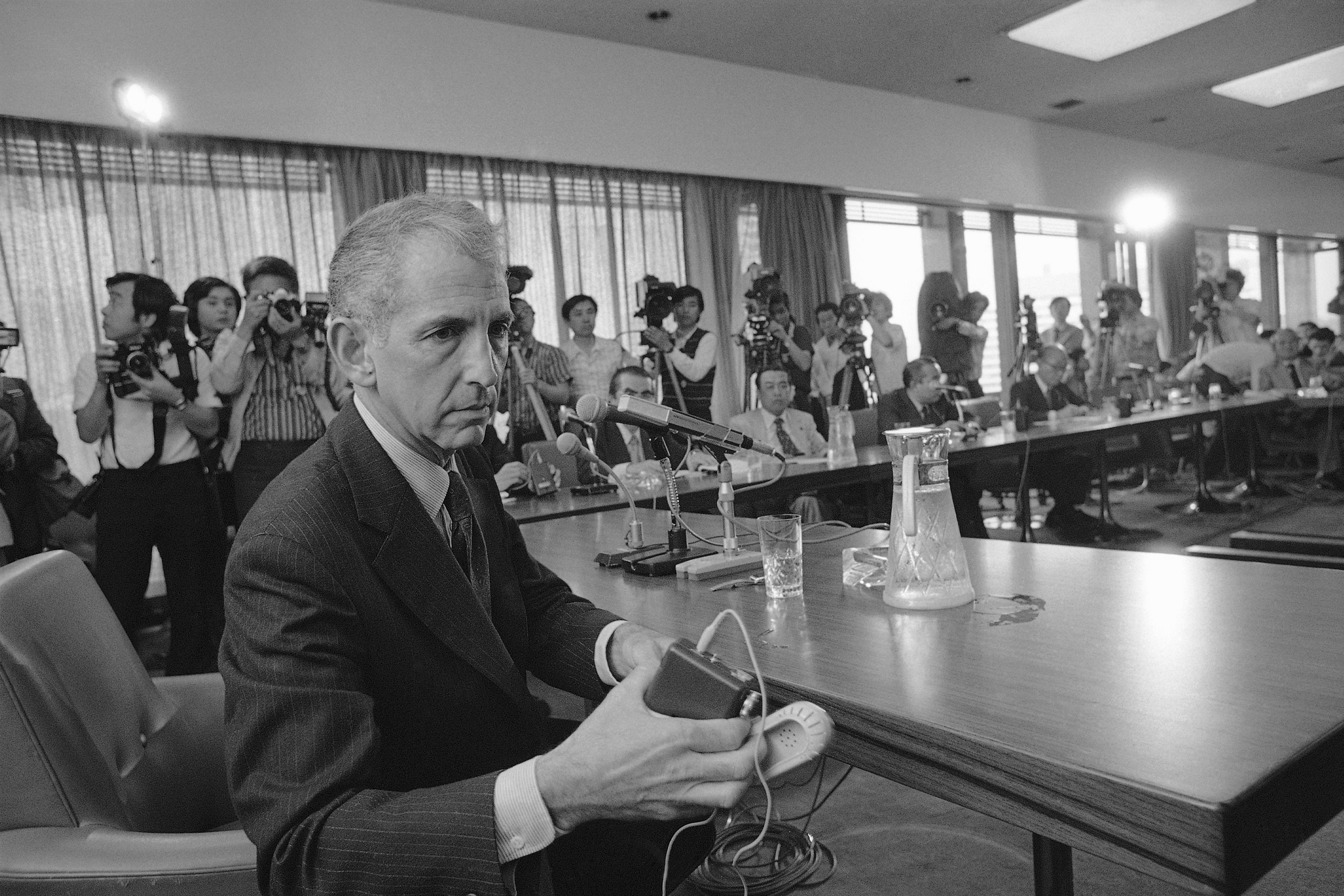 Ellsberg, hair gray, is dressed in a suit, and looking to one side as he sits at a long wooden table with microphones, and holds what looks like a Sony Walkman. Photo is black and white. Along one wall are Japanese photographers, snapping photos and filming him. 