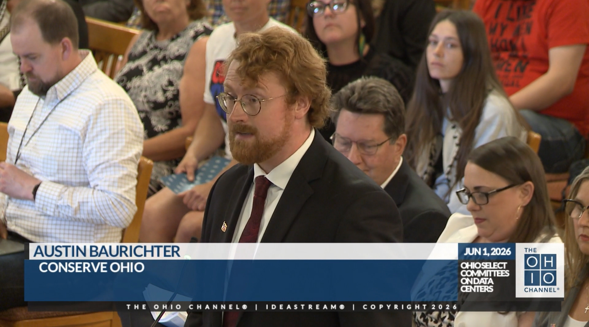 A man in a black suit speaks in front of the Ohio General Assembly’s Select Committee on Data Centers while seated spectators look on in the background.