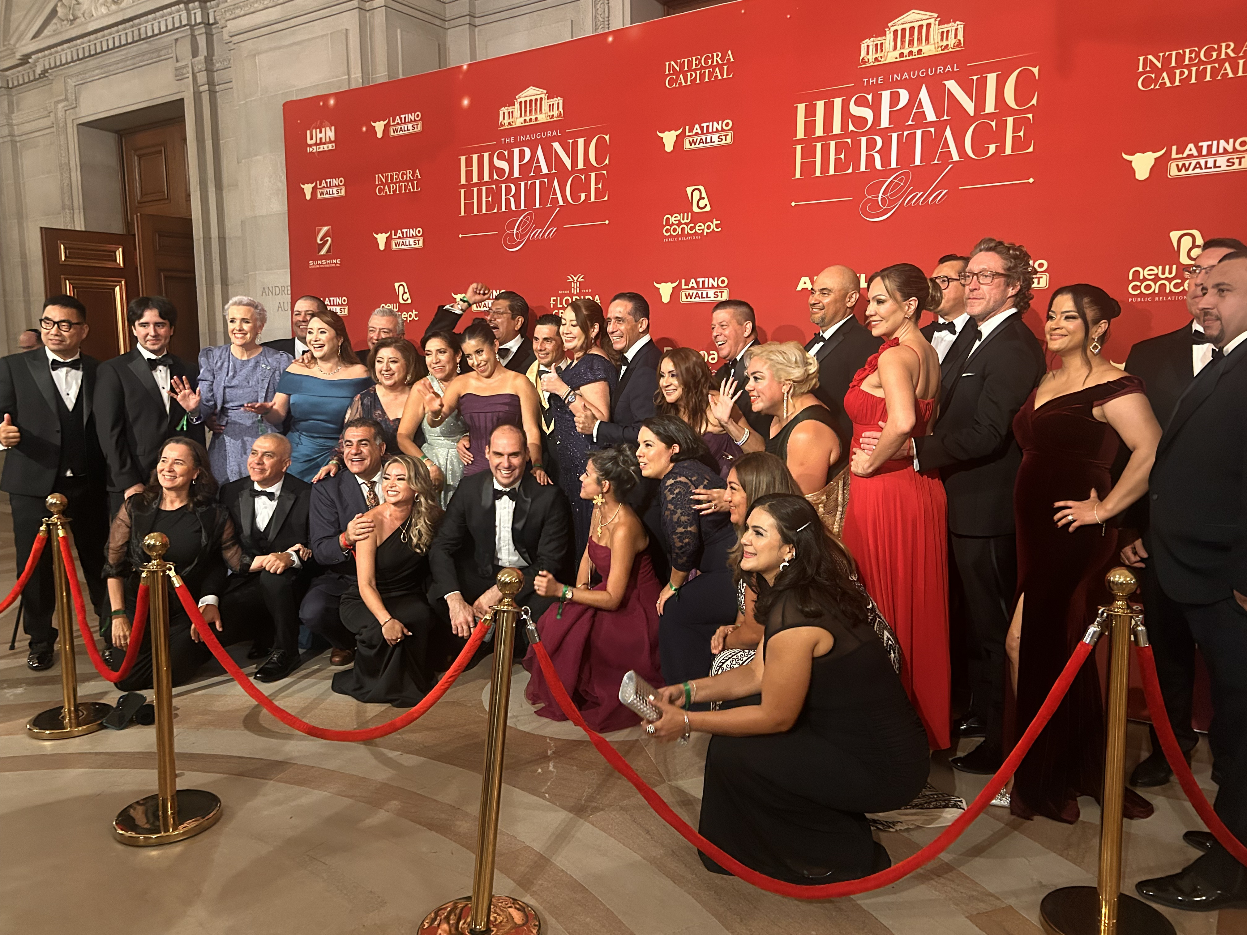 A group of people pose for a photo in front of a red wall reading Hispanic Heritage Gala.