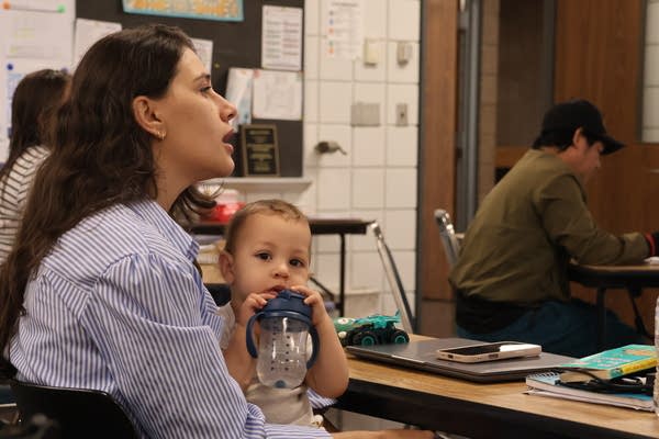 A baby teeths on a bottle while being held by his mother, who sits at a classroom desk.