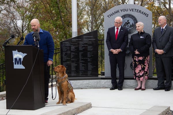 A man speaking at a podium with a dog sitting by him