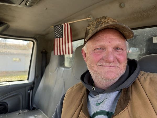 A man sits in a truck with a small American flag hanging behind him.