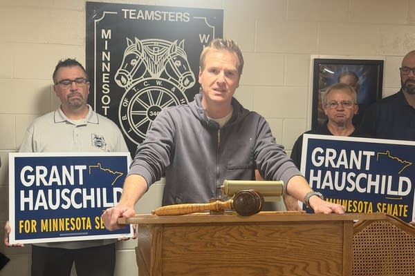 A man speaks from behind a podium while people behind him hold political signs.