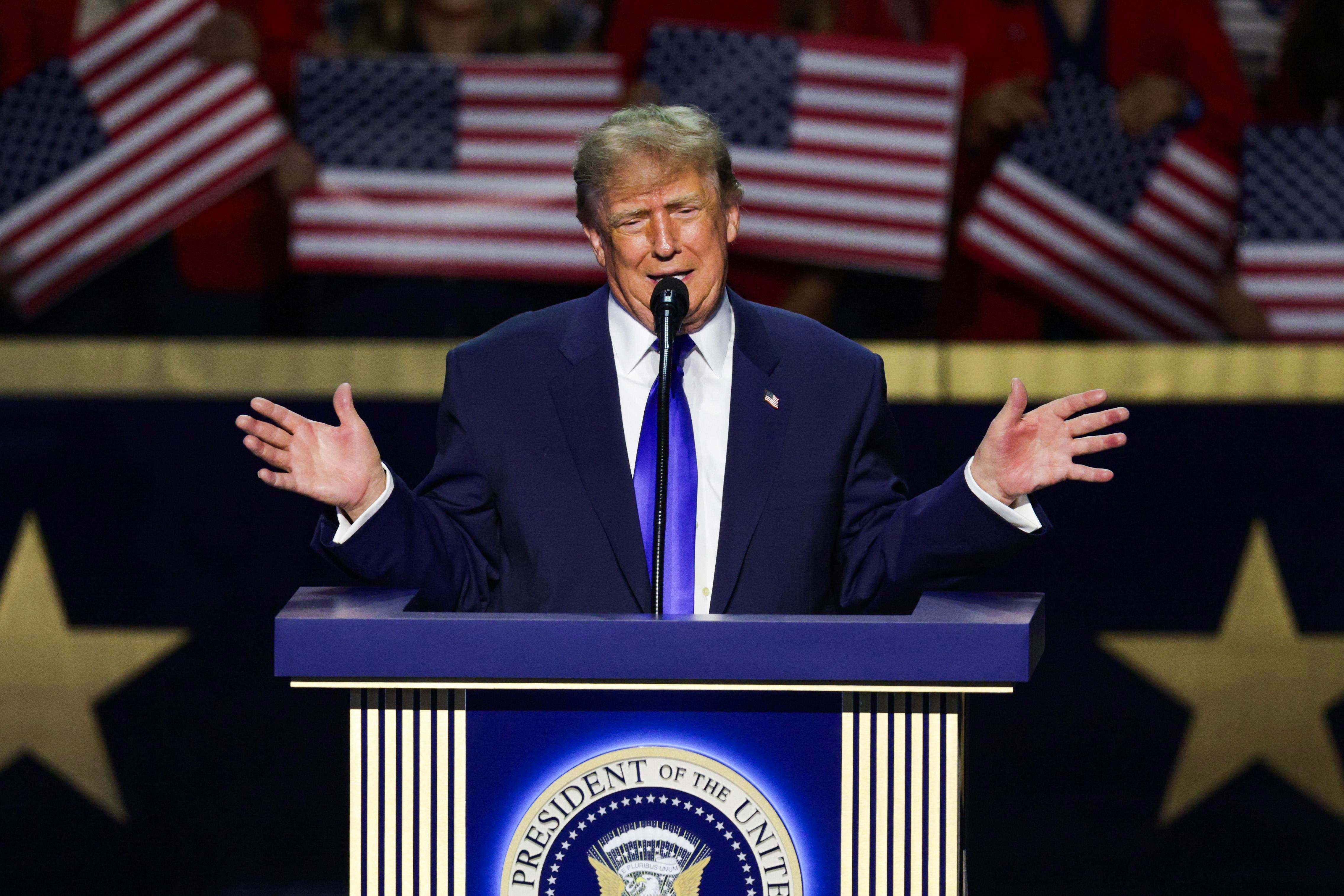 Donald Trump speaks on stage against a background of American flags.