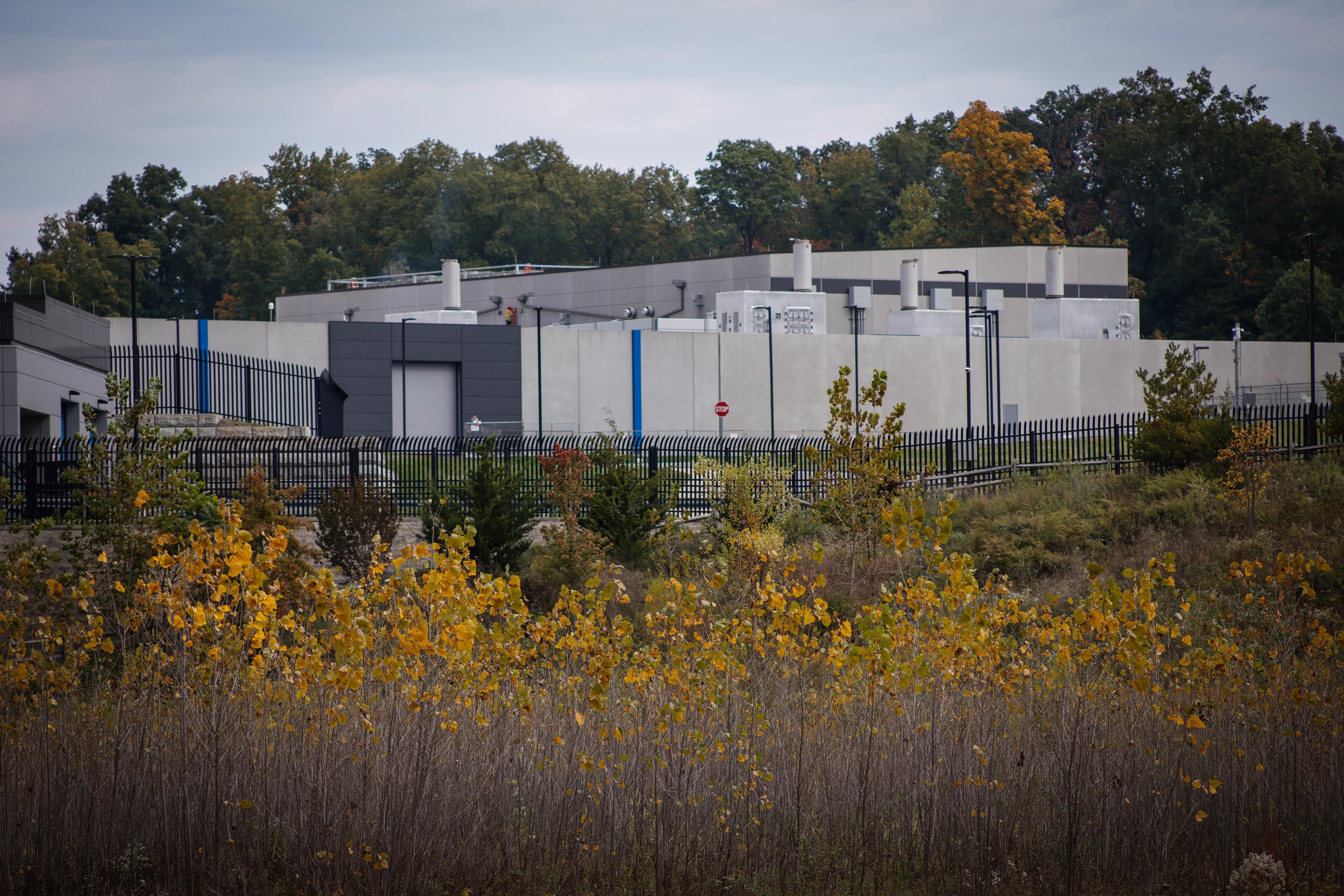 A data center in shades of gray, surrounded by a fence, is seen in front of trees beginning to show autumnal colors; in the foreground is more autumnal greenery.