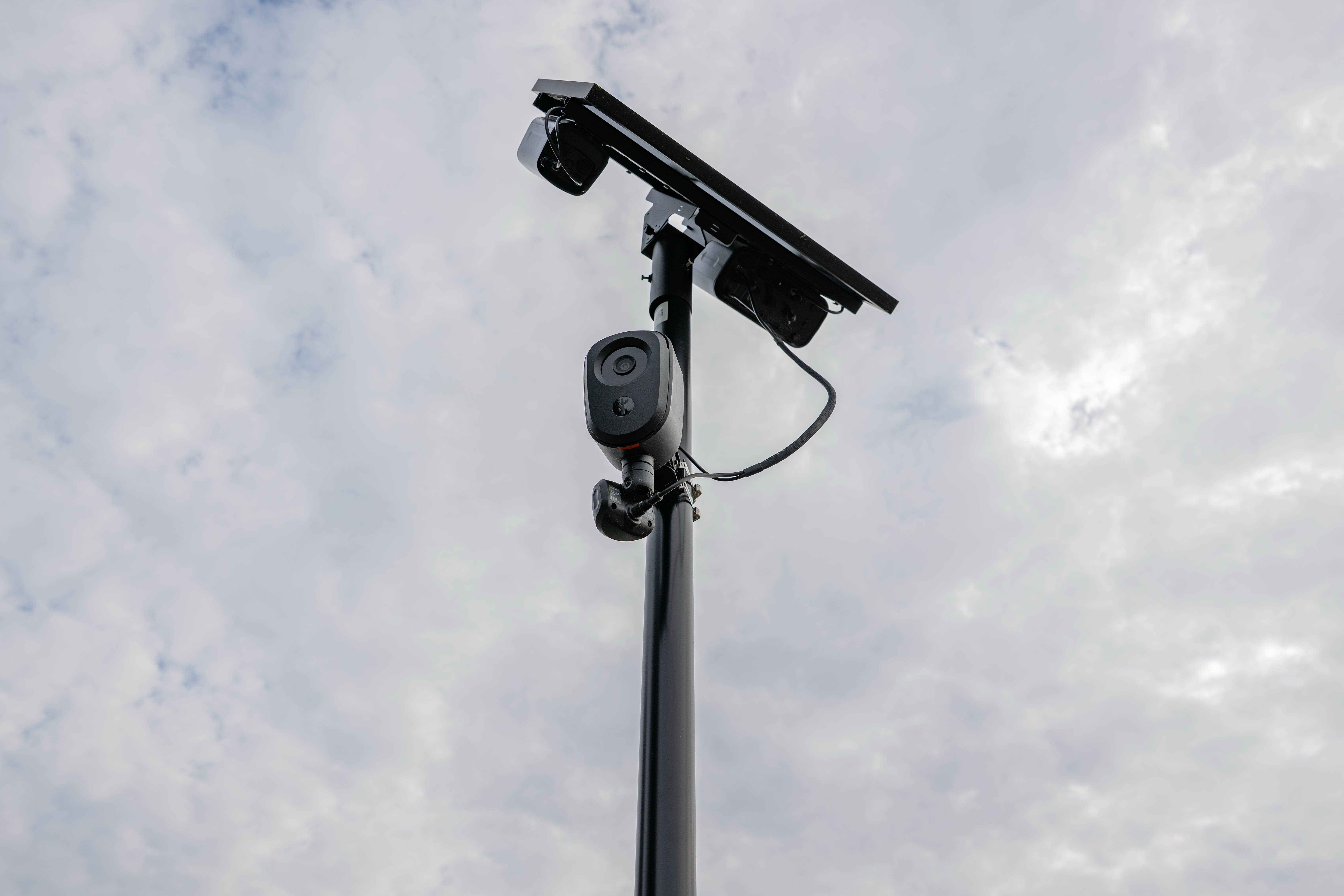 A Flock camera attached to a light post, beneath a solar panel, on a cloudy day.
