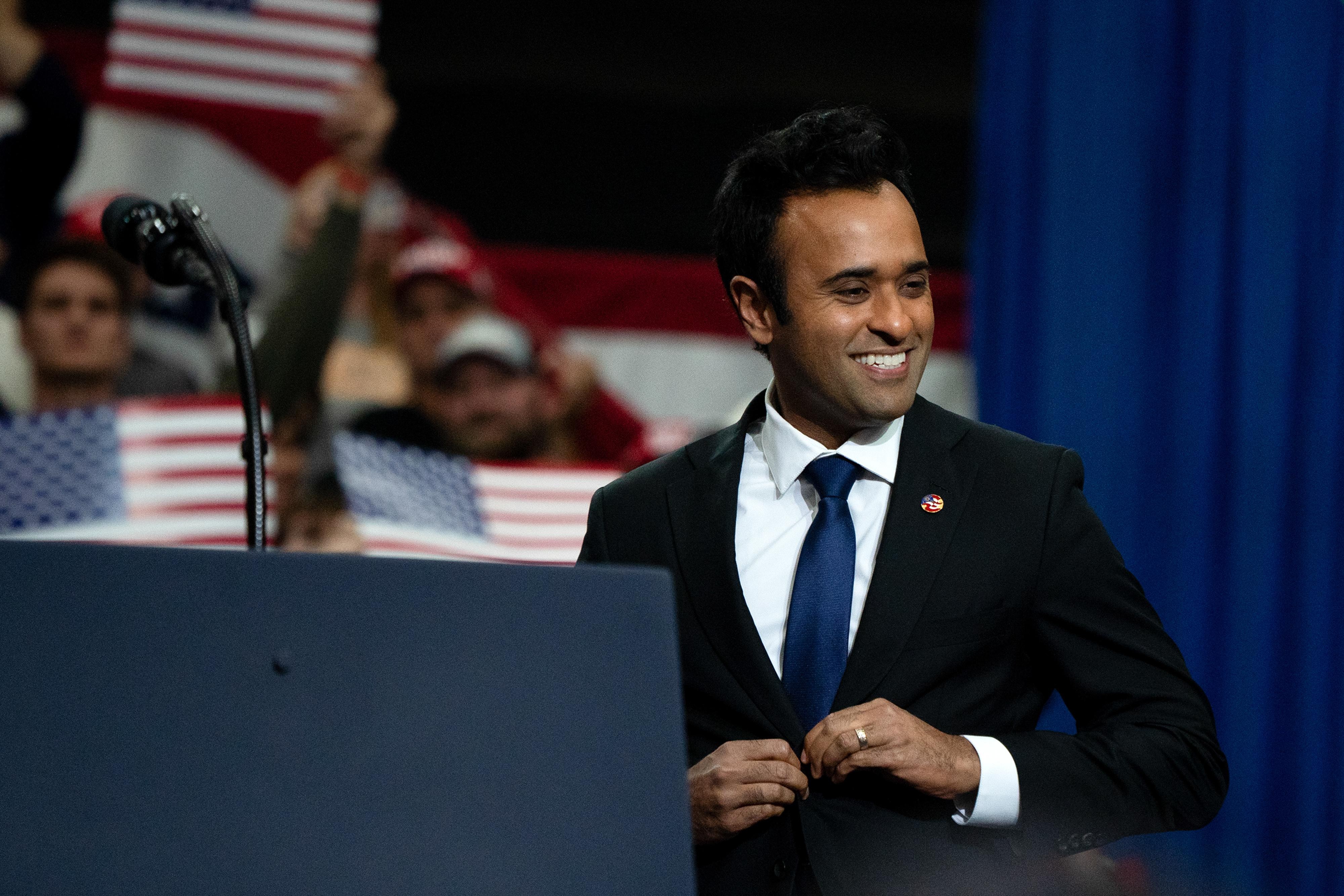 Vivek Ramaswamy, wearing a suit and tie, walks up to a podium on stage; behind him, supporters hold American flag signs.